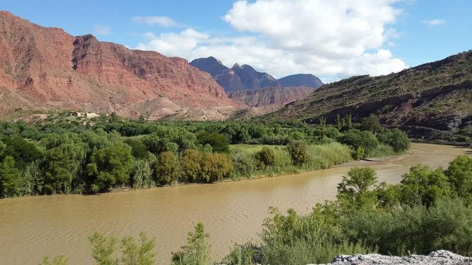Un río bordea vegetación verde y cerros rojizos bajo un cielo azul con nubes.