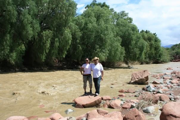 Dos personas están de pie sobre una roca junto a un río y árboles frondosos.