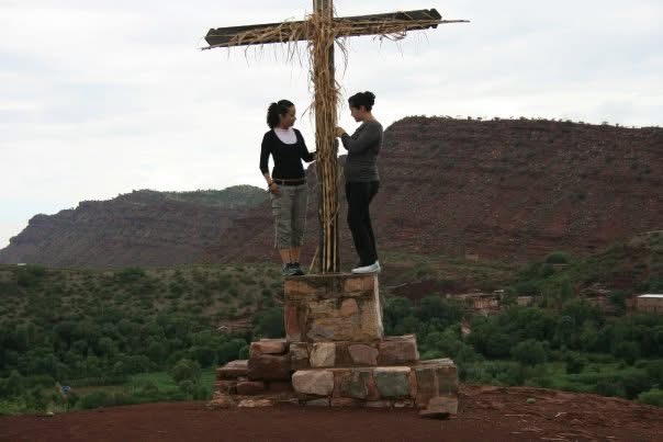 Dos personas junto a una cruz de madera sobre una estructura de piedra, con cerros al fondo.